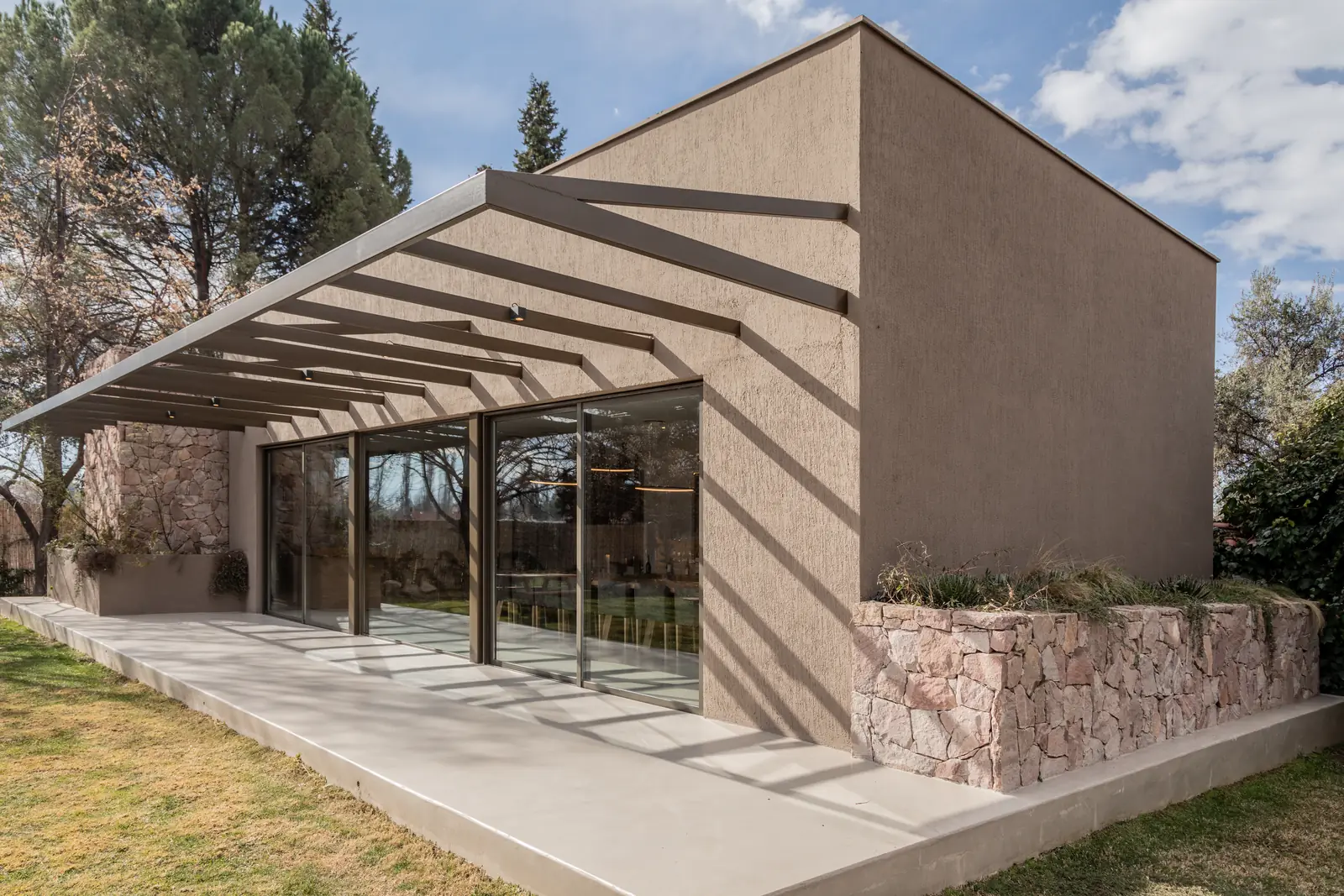 Vista lateral completa con pérgola, carpintería vidriada y muro de piedra al pie