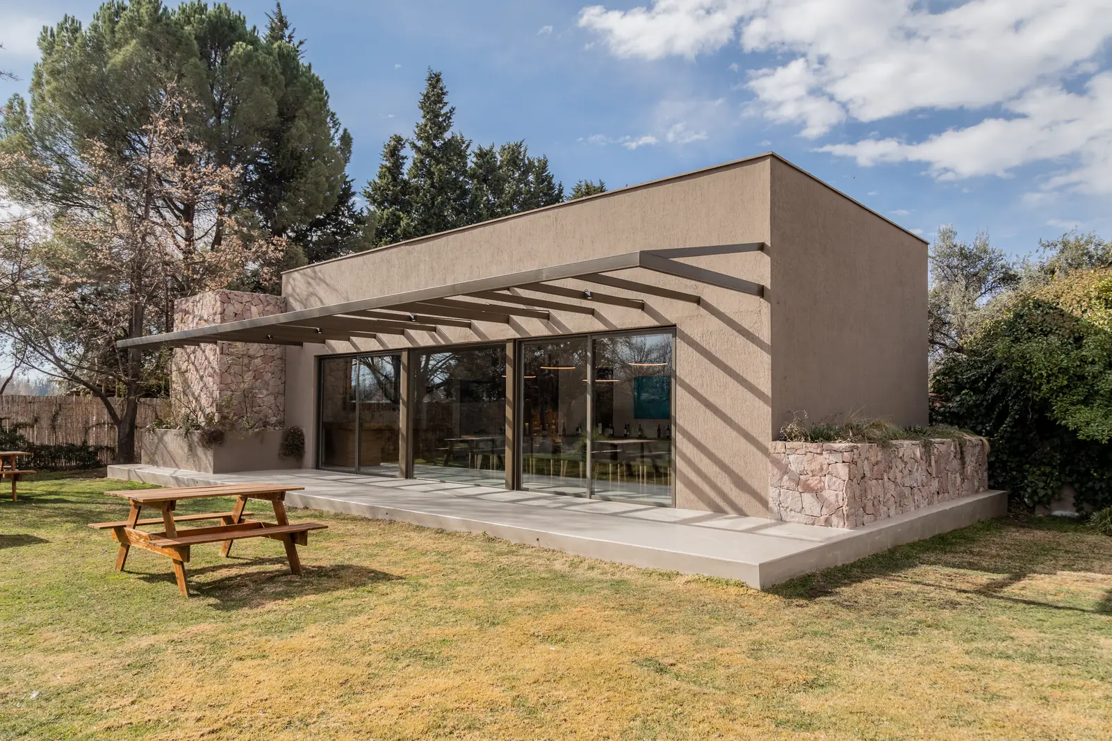 Exterior con pérgola de perfiles metálicos, cerramiento vidriado y muro de piedra bajo