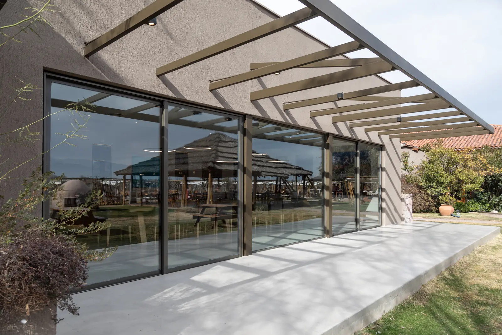 Galería vidriada bajo pérgola inclinada, con reflejos del parque en los cristales