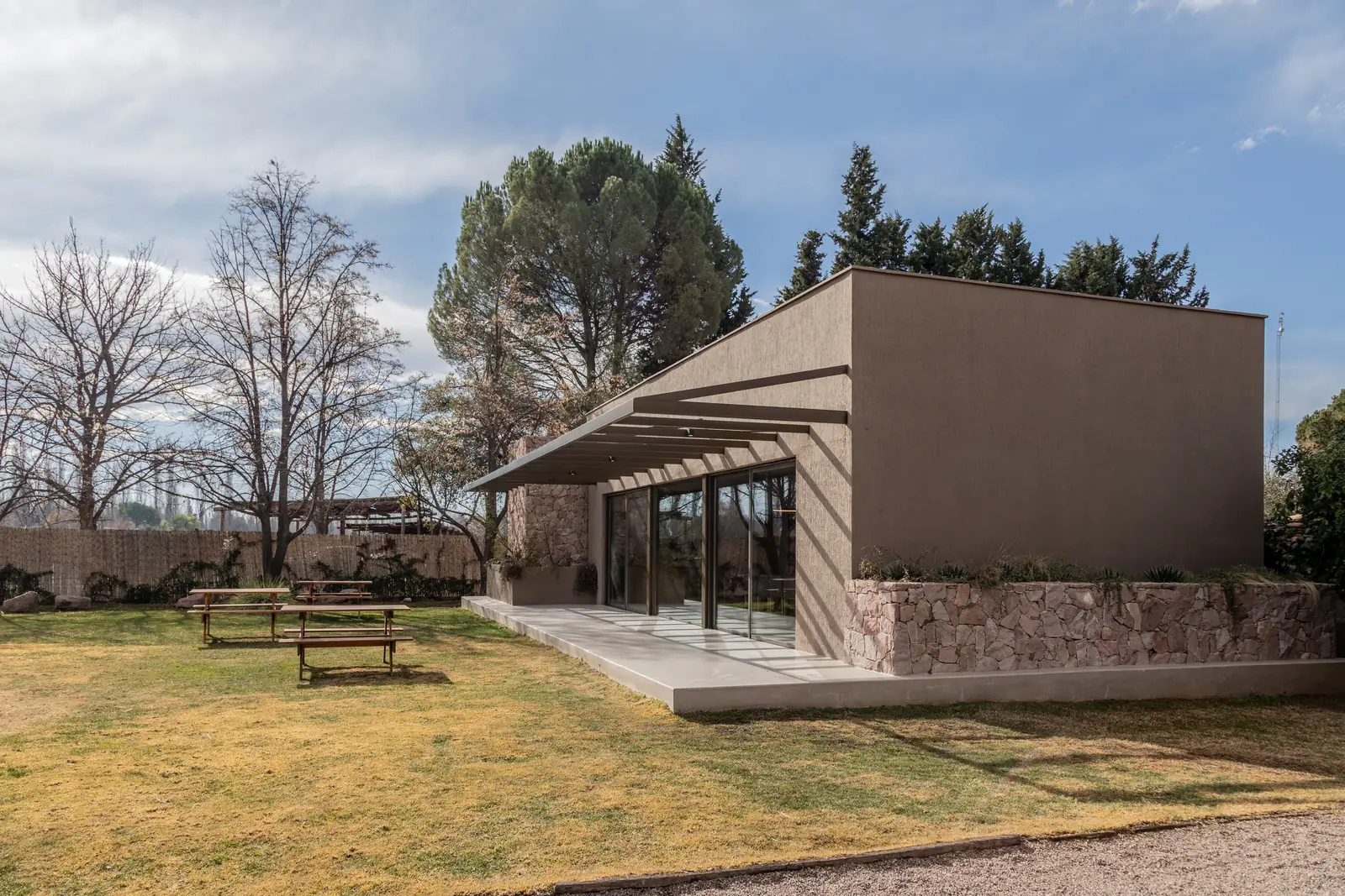 Volumen de la sala recortado entre el parque, con pérgola y mesas de picnic