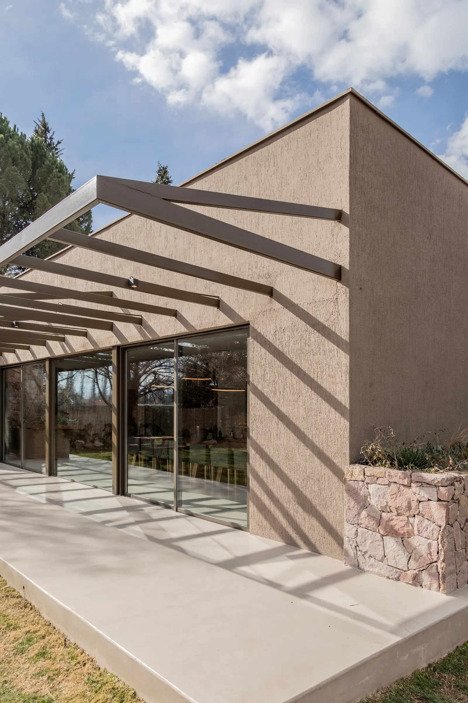 Esquina del volumen revocado con pérgola metálica y zócalo de piedra vista