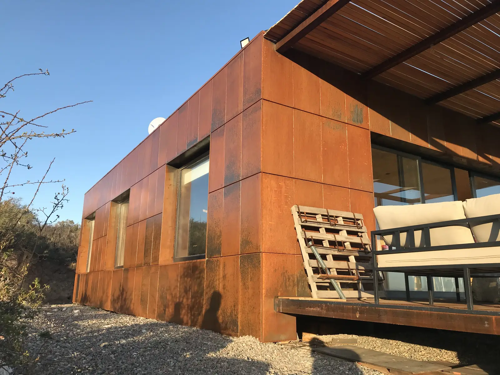 Fachada de chapas de acero corten con ventanas alargadas y banco de madera