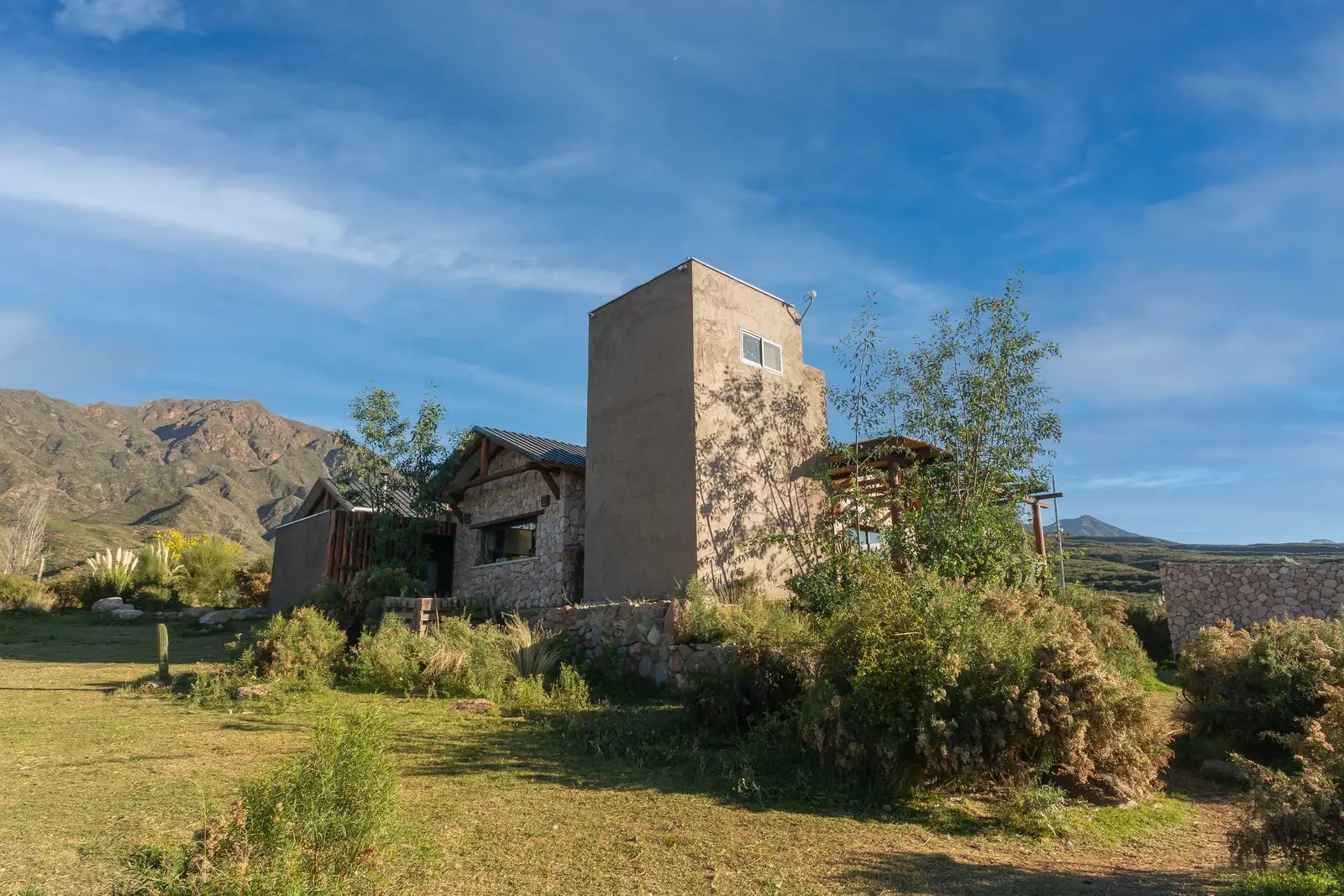 Casa de campo con torre revocada y montañas al fondo, entre jardín seco