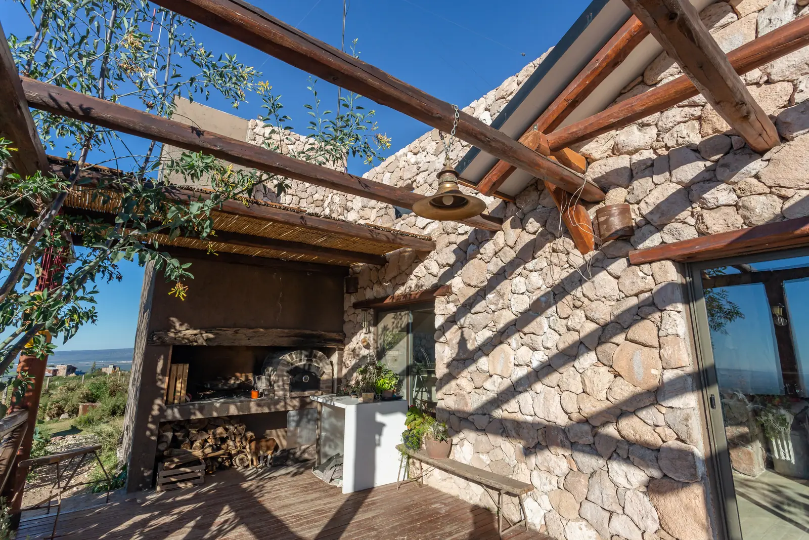 Quincho abierto con parrilla, pérgola de troncos y muro de piedra al sol
