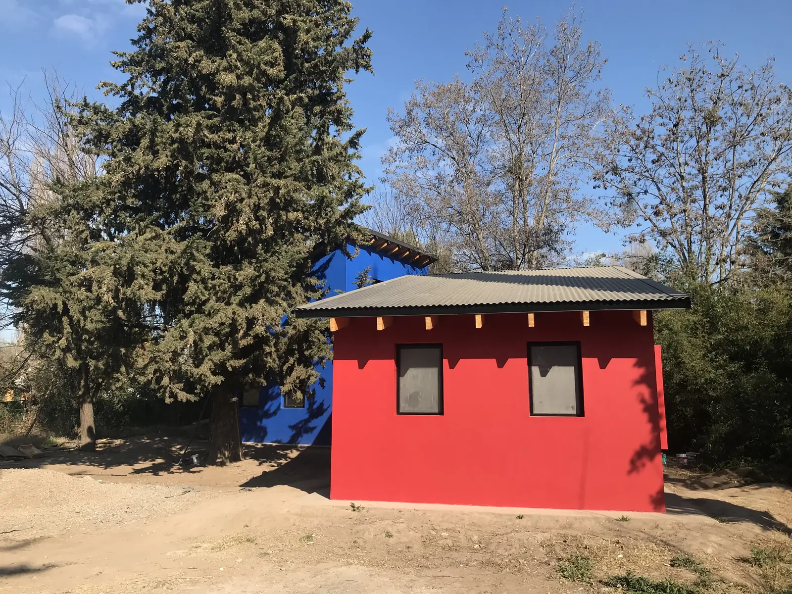 Casita roja y volumen azul contiguo vistos desde el camino de tierra