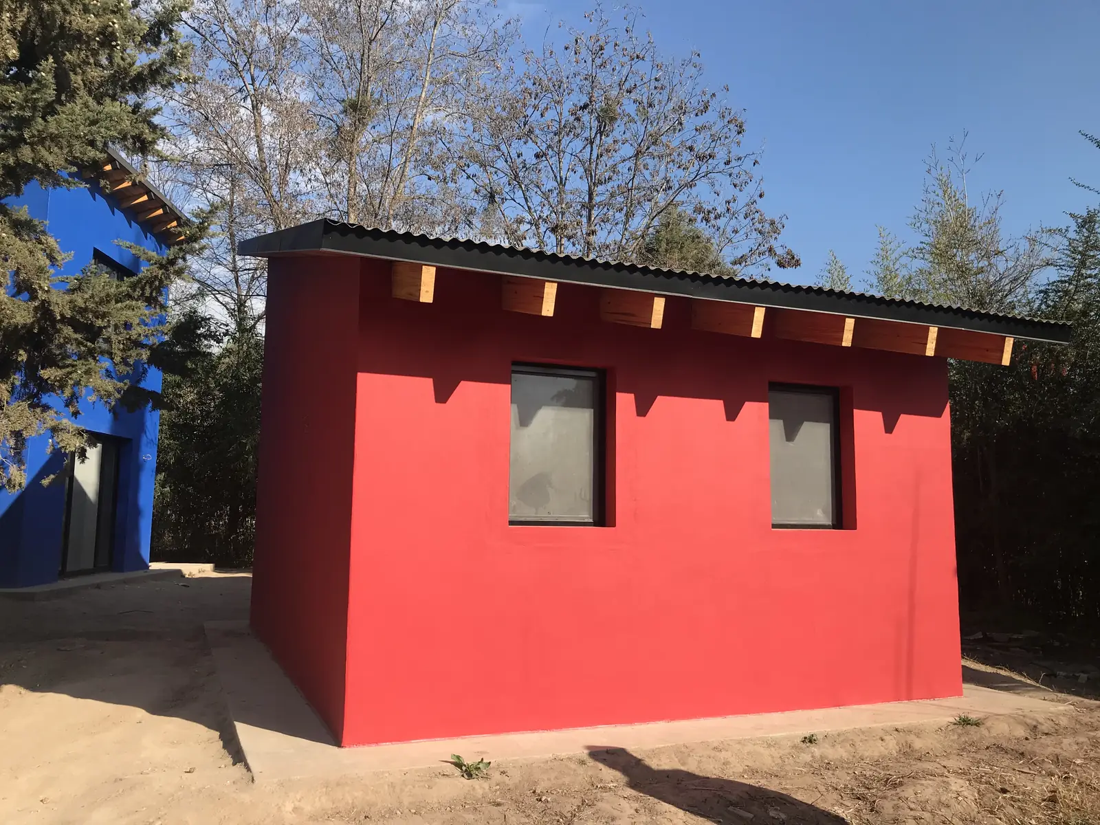 Casita roja con techo a un agua y ventanas entre árboles añosos