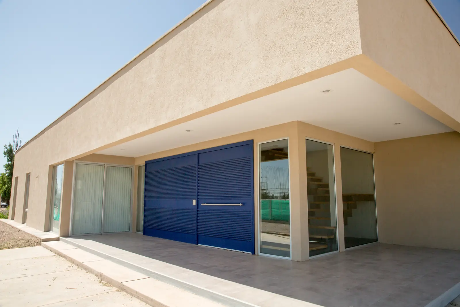 Galería cubierta bajo alero profundo con portón azul y cerramientos vidriados