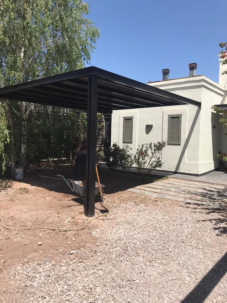 Cochera con estructura metálica negra junto a la vivienda terminada y el jardín