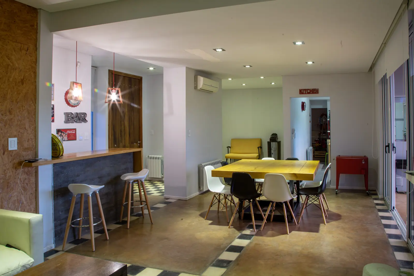 Estar comedor integrado con barra, banquetas y mesa amarilla bajo luces colgantes