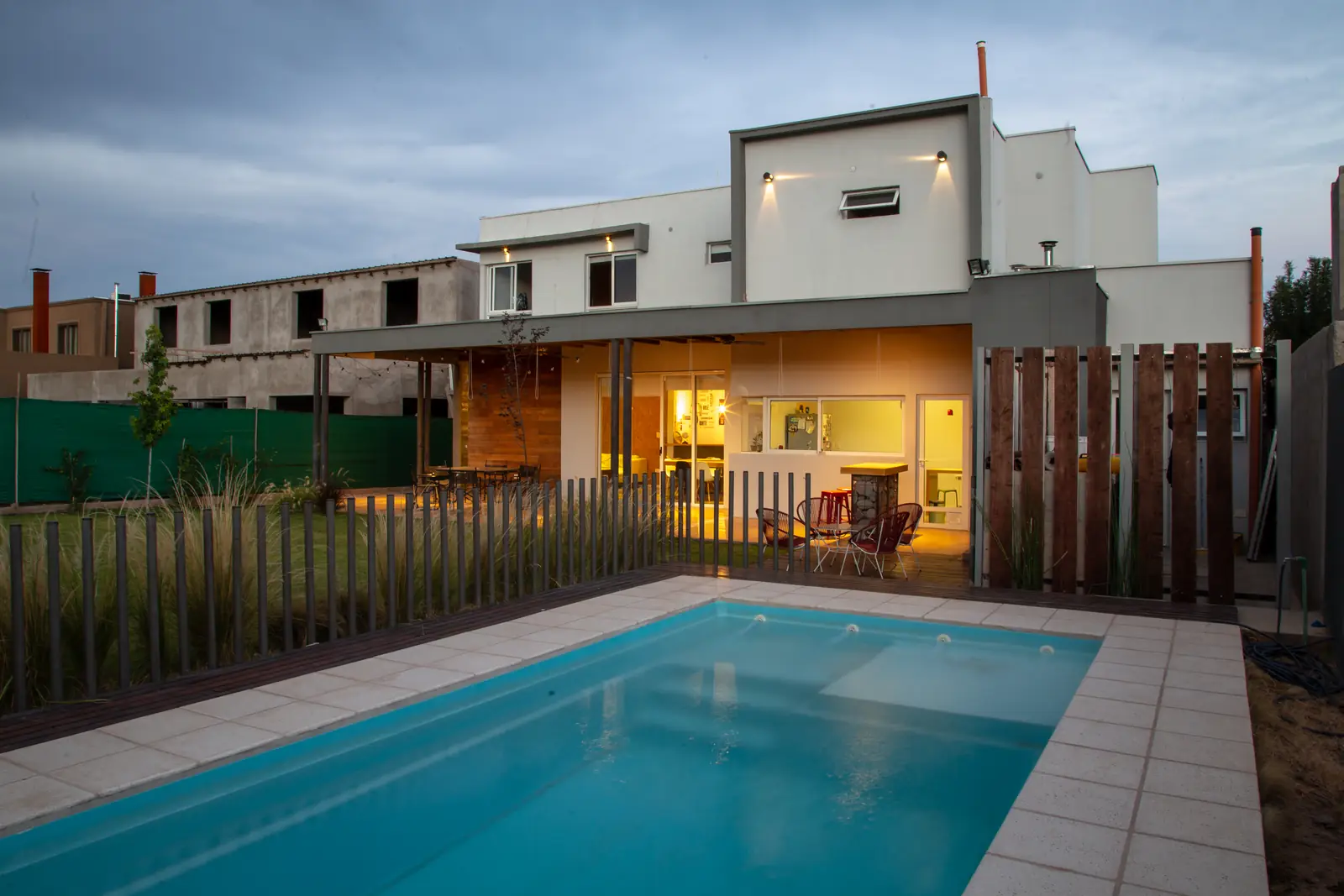 Pileta rectangular con borde de baldosas, cerco de madera y la casa iluminada detrás