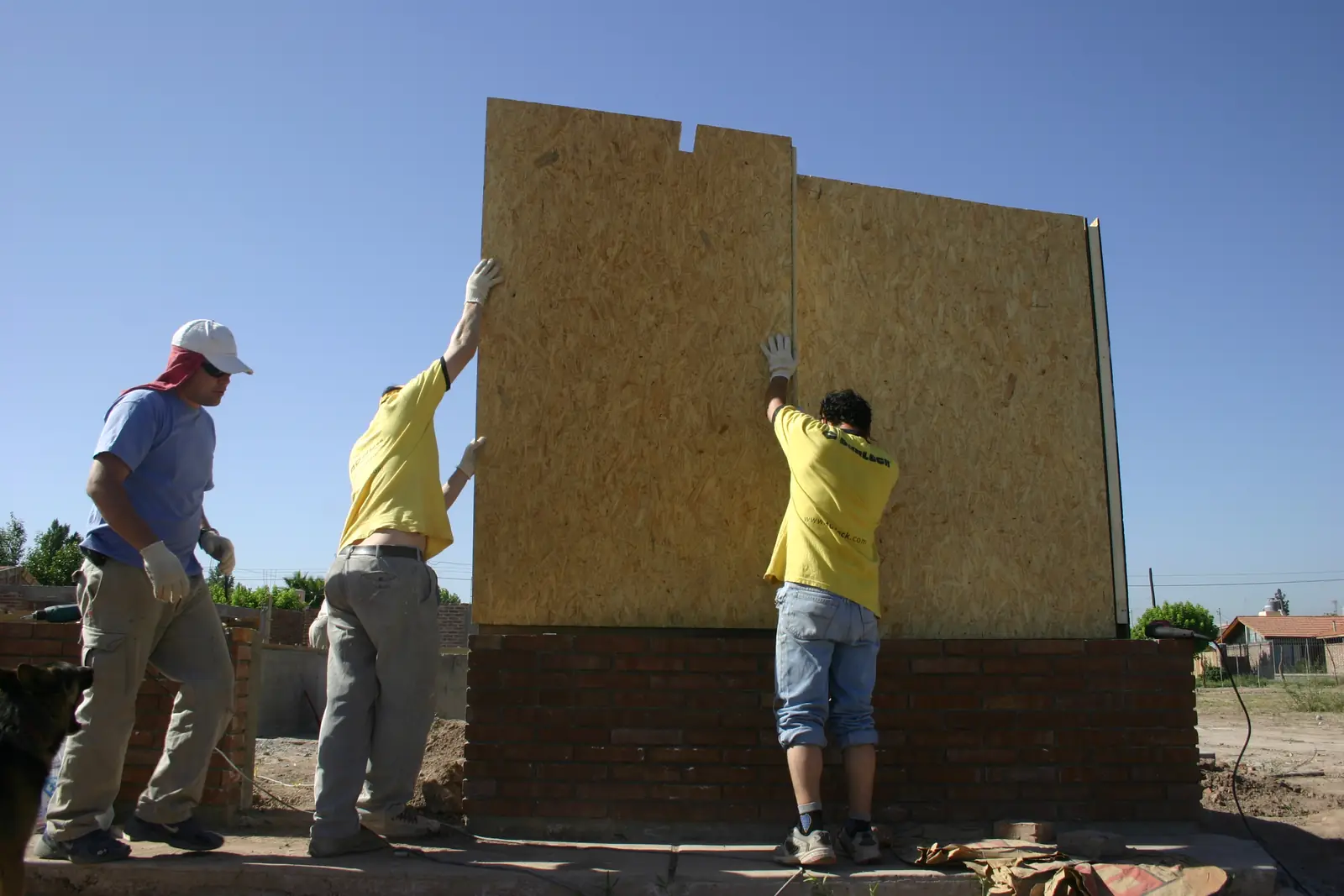 Tres operarios levantan un panel SIP sobre el muro de ladrillo de base
